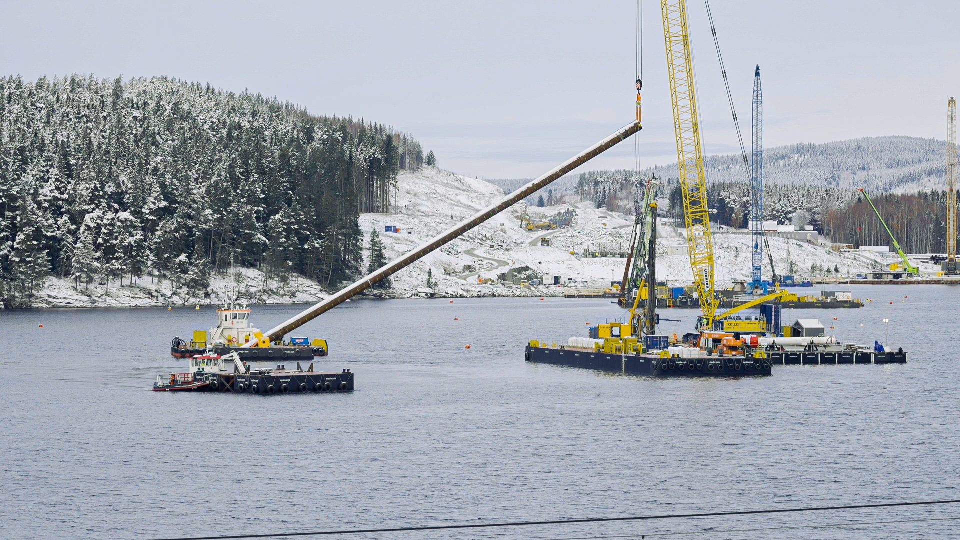 Crane barge lifting a long pile on a lake below a snow-covered forest