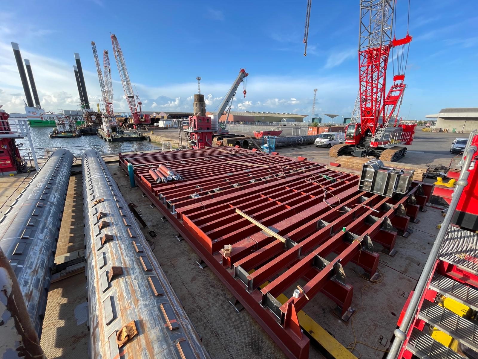 Red steel grillage laid out on a barge deck beside tubular piles, with cranes on the quay