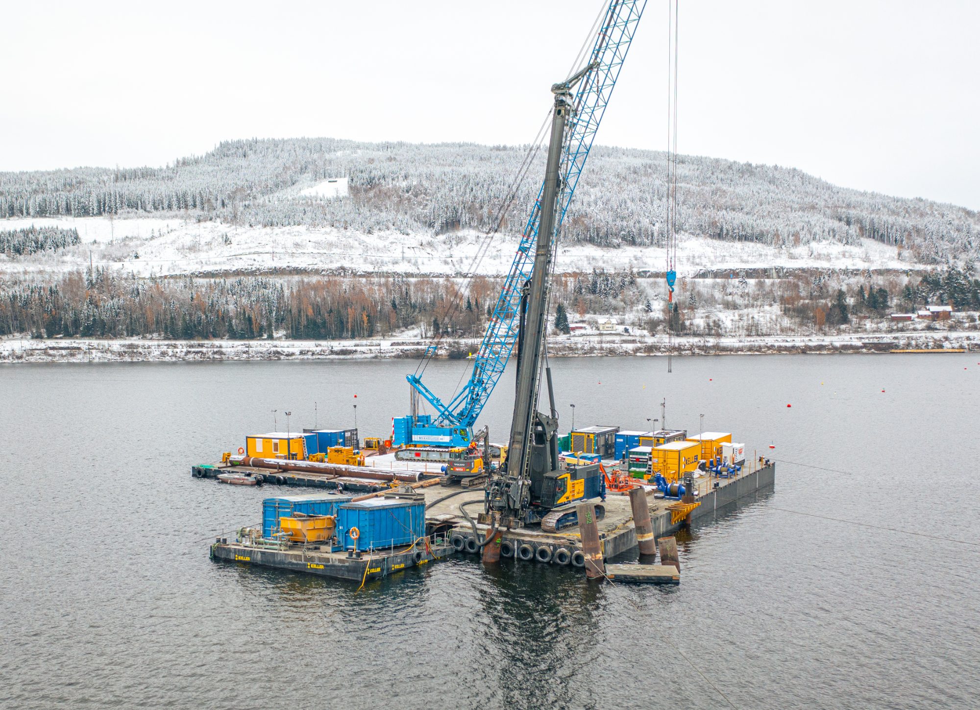 Crane barge with a piling rig working on a lake beneath a snow-covered hillside