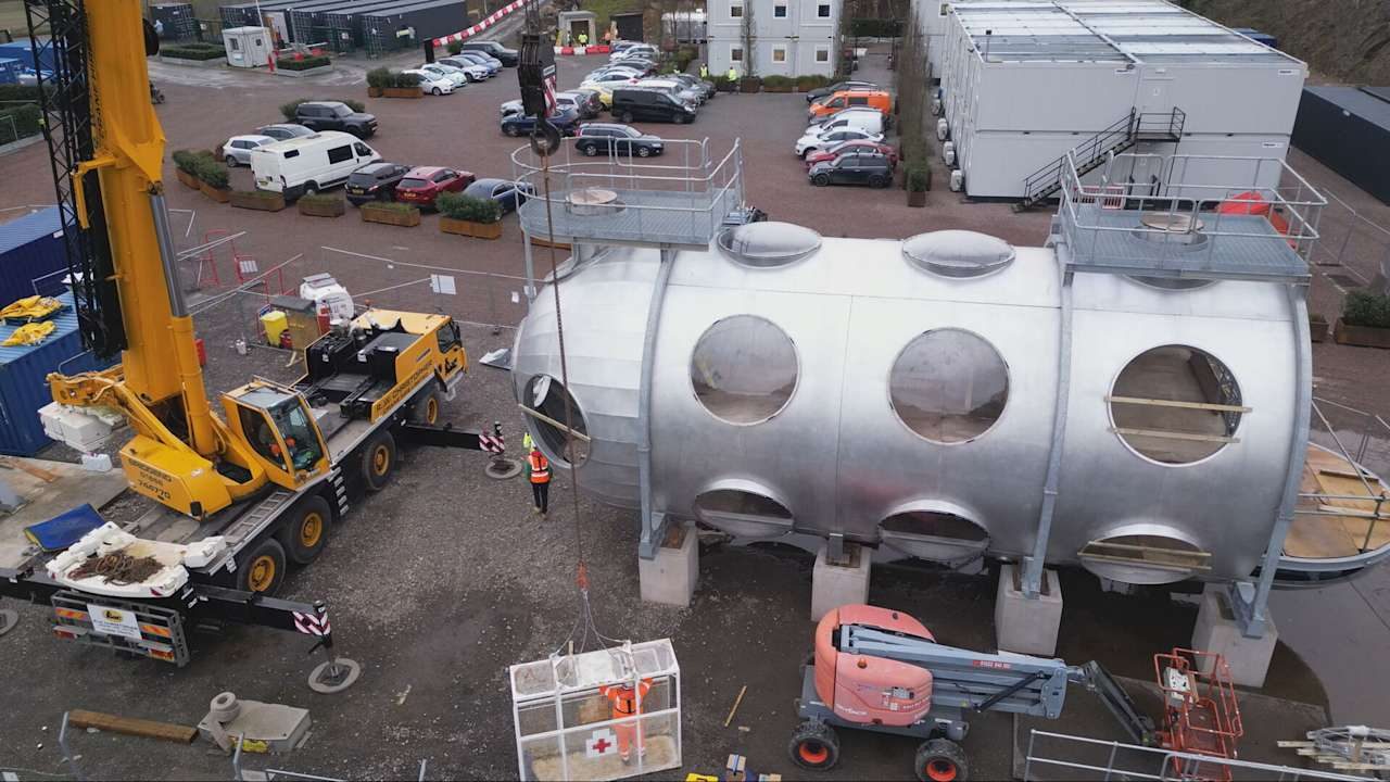 Mobile crane positioning a large cylindrical chamber with circular hatches on a construction site