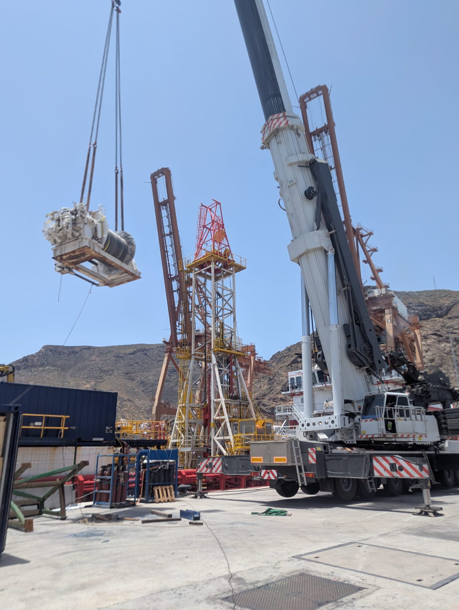 Mobile crane lifting a winch package beside a drilling derrick on a quayside