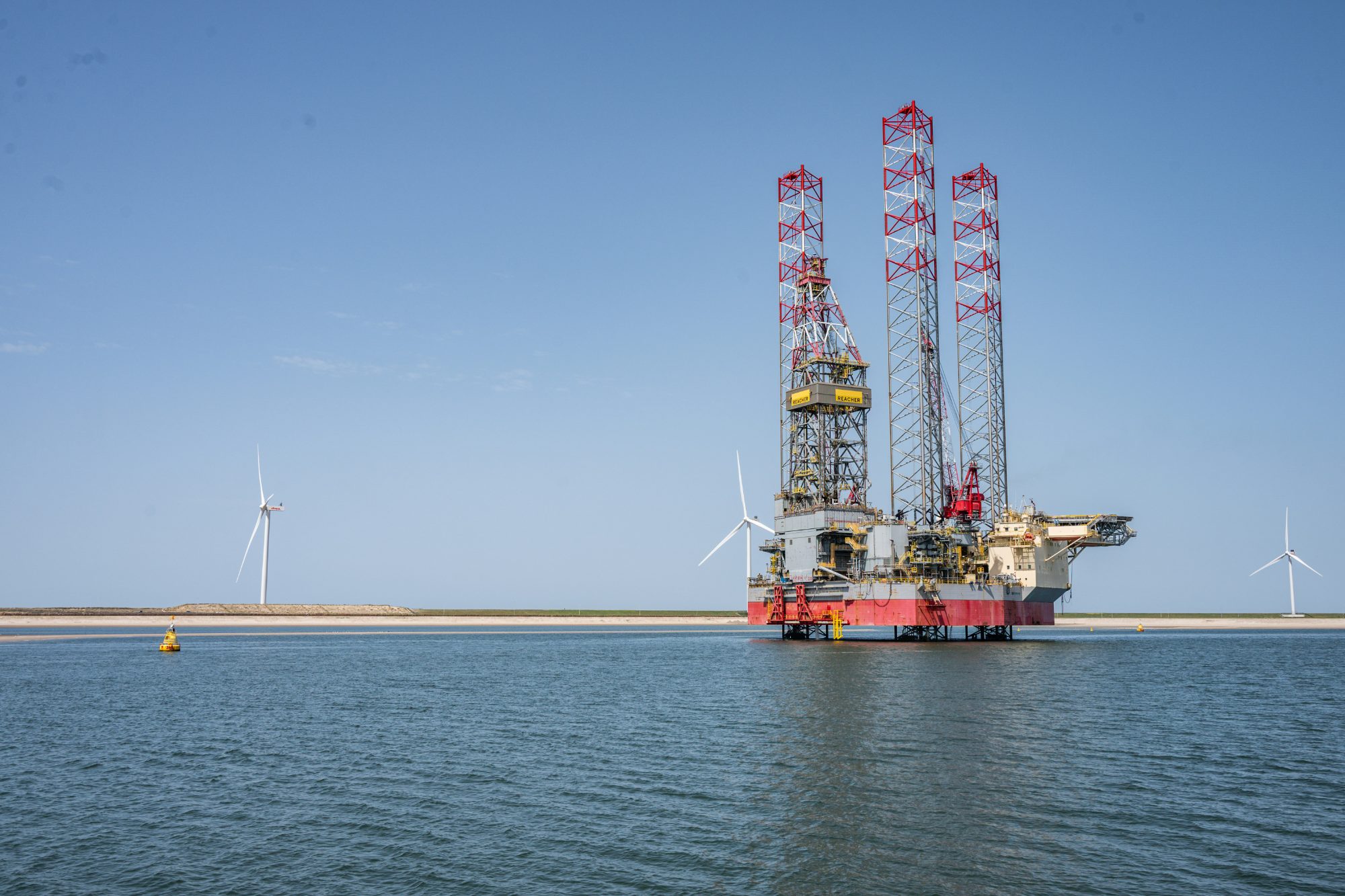 Jack-up drilling rig standing in shallow water with wind turbines on the shore behind