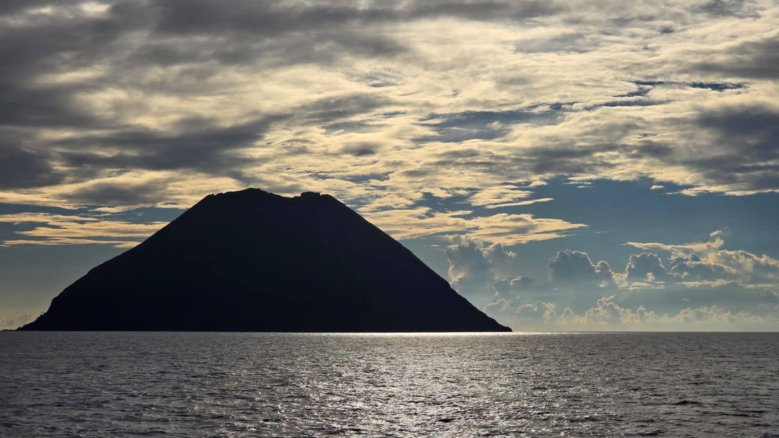 Conical island silhouetted against a cloudy sky over calm water