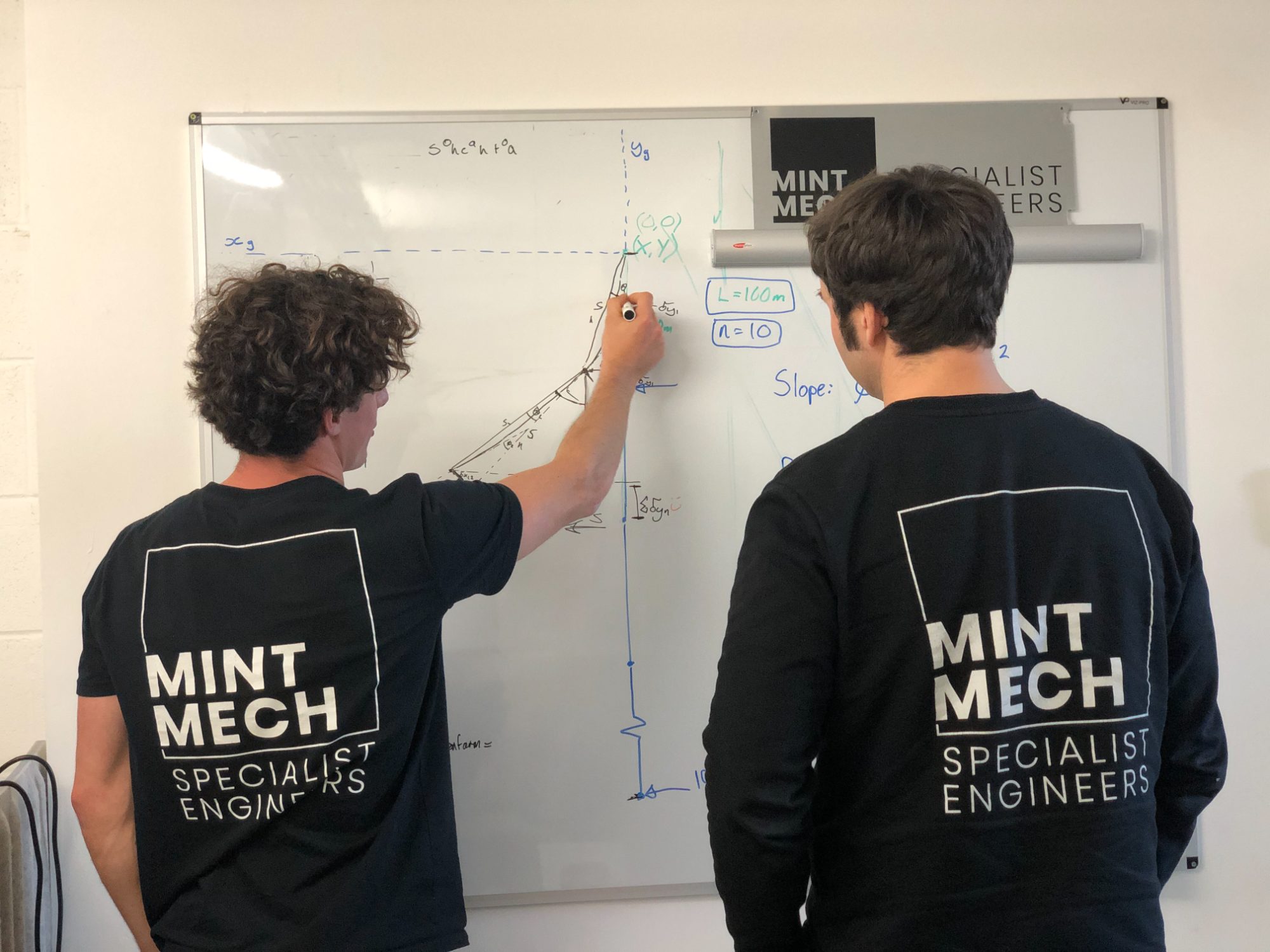 Two engineers in MintMech shirts working through a sketch on a whiteboard