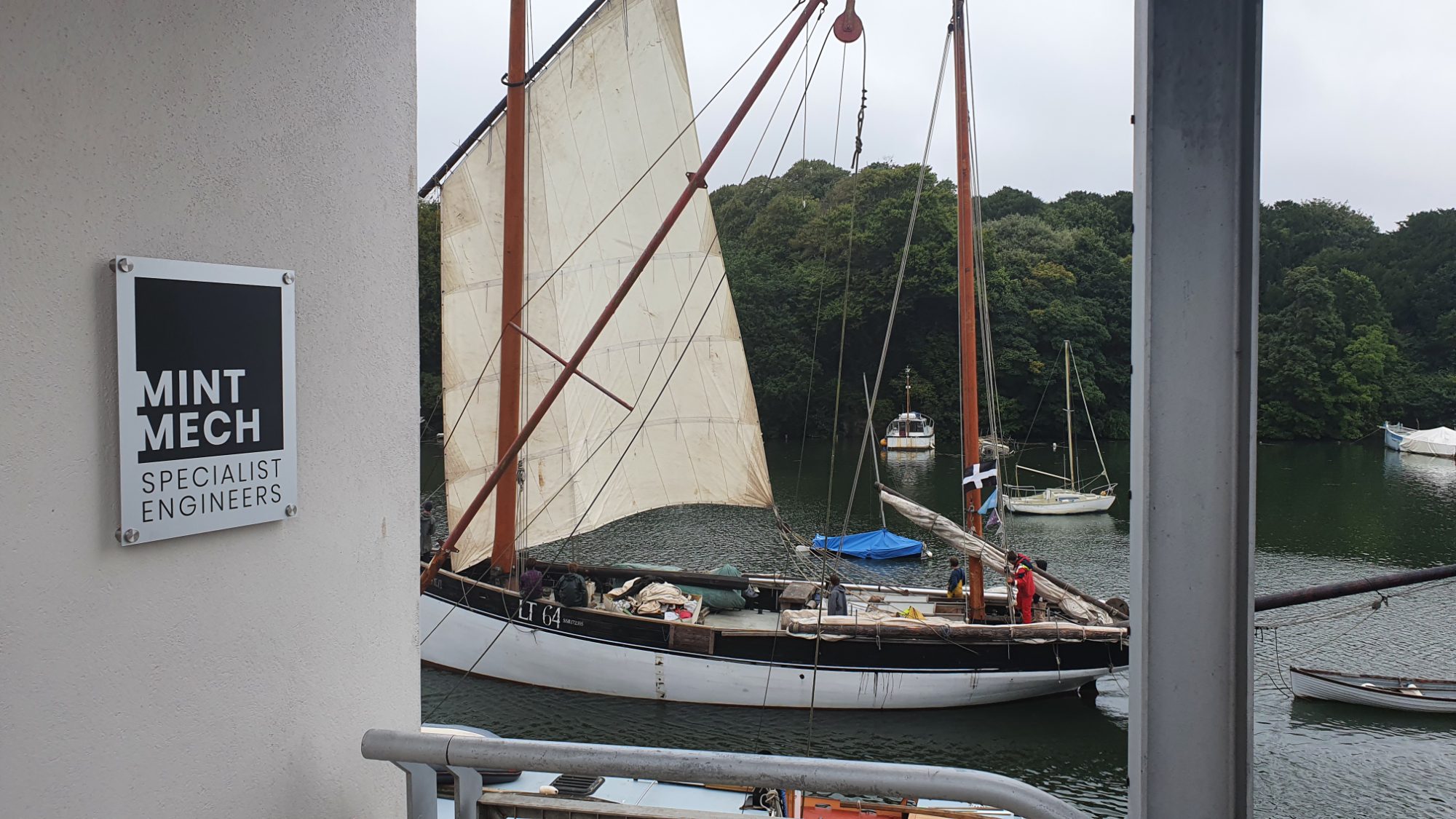 MintMech sign on a quayside wall with a traditional sailing boat moored behind
