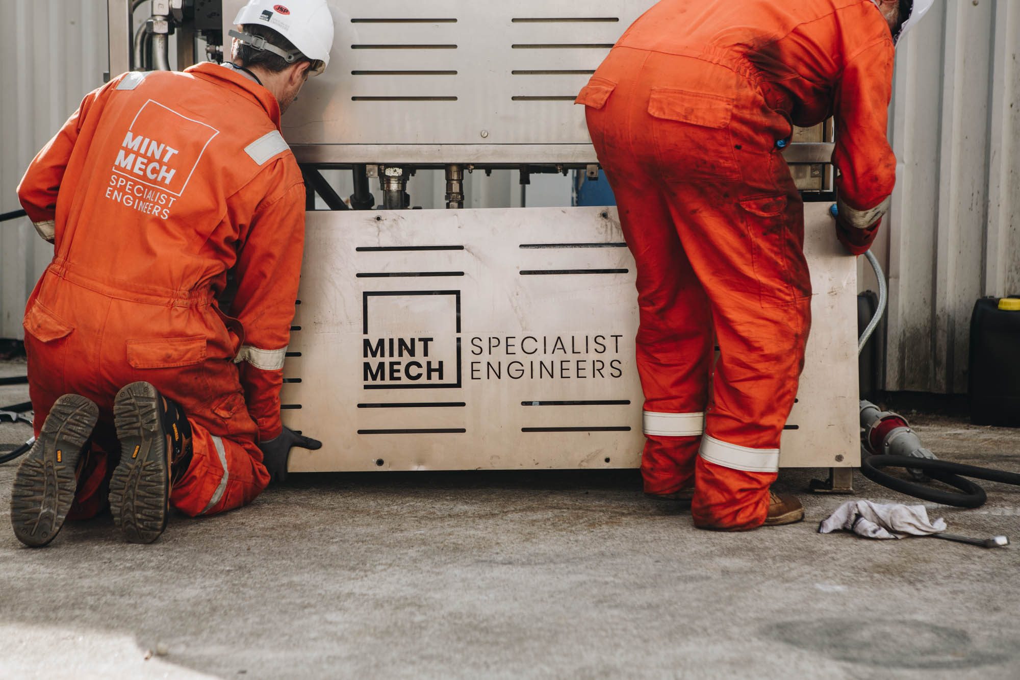 Two engineers in orange coveralls working on a MintMech equipment cabinet