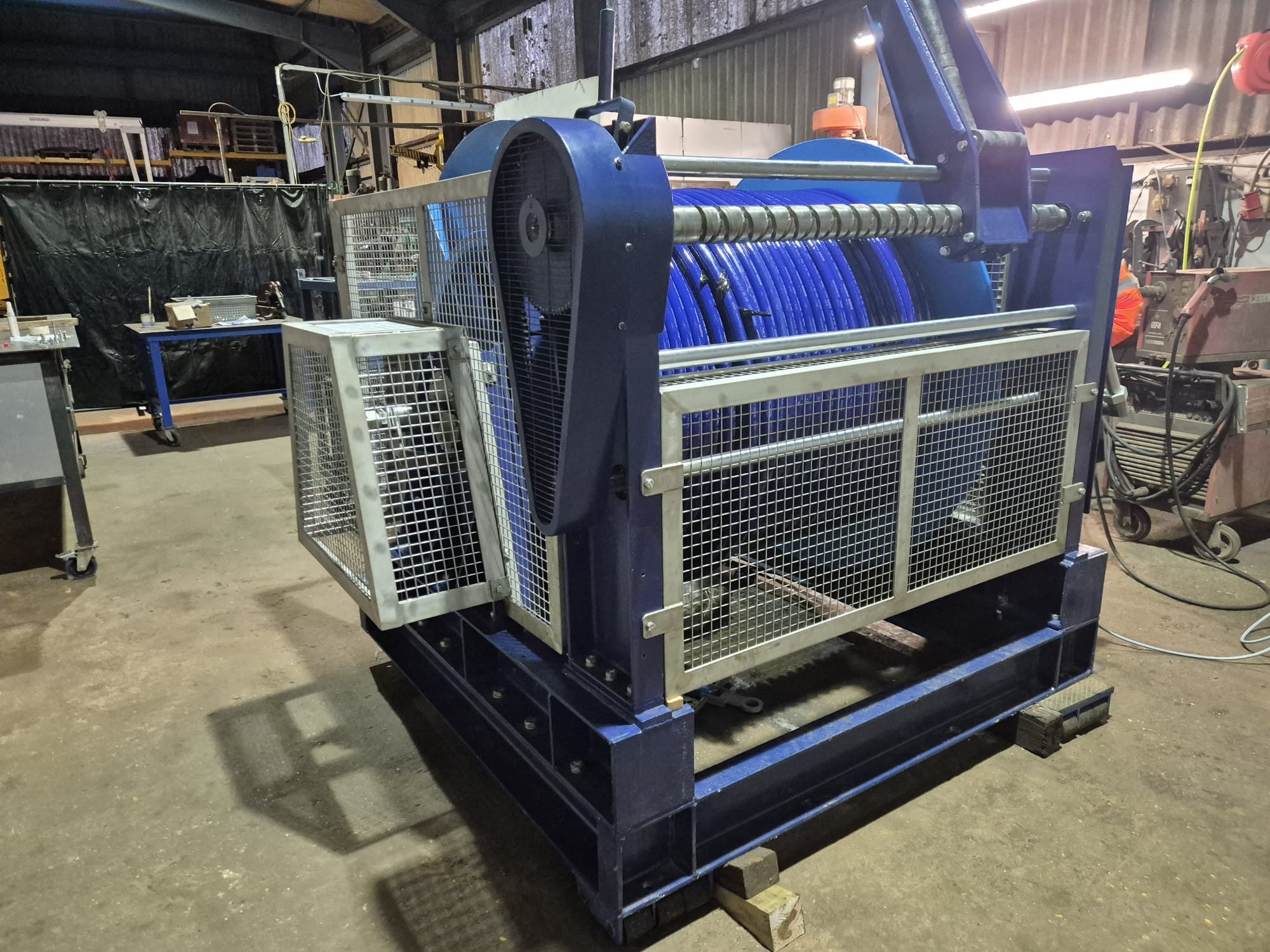 Blue winch with a spooling drum behind a galvanised mesh guard in a workshop