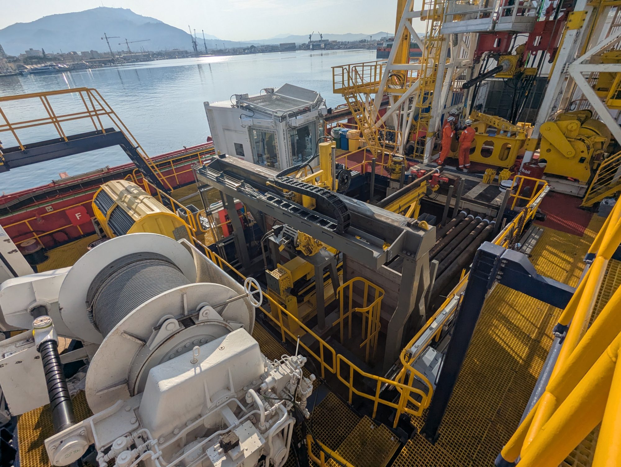 View across a drilling vessel's deck showing a winch, pipe handling equipment and yellow walkways