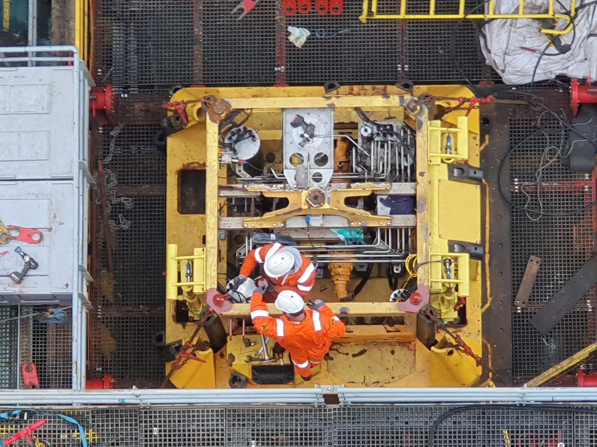 Two engineers in orange coveralls working on yellow deployment equipment, seen from above