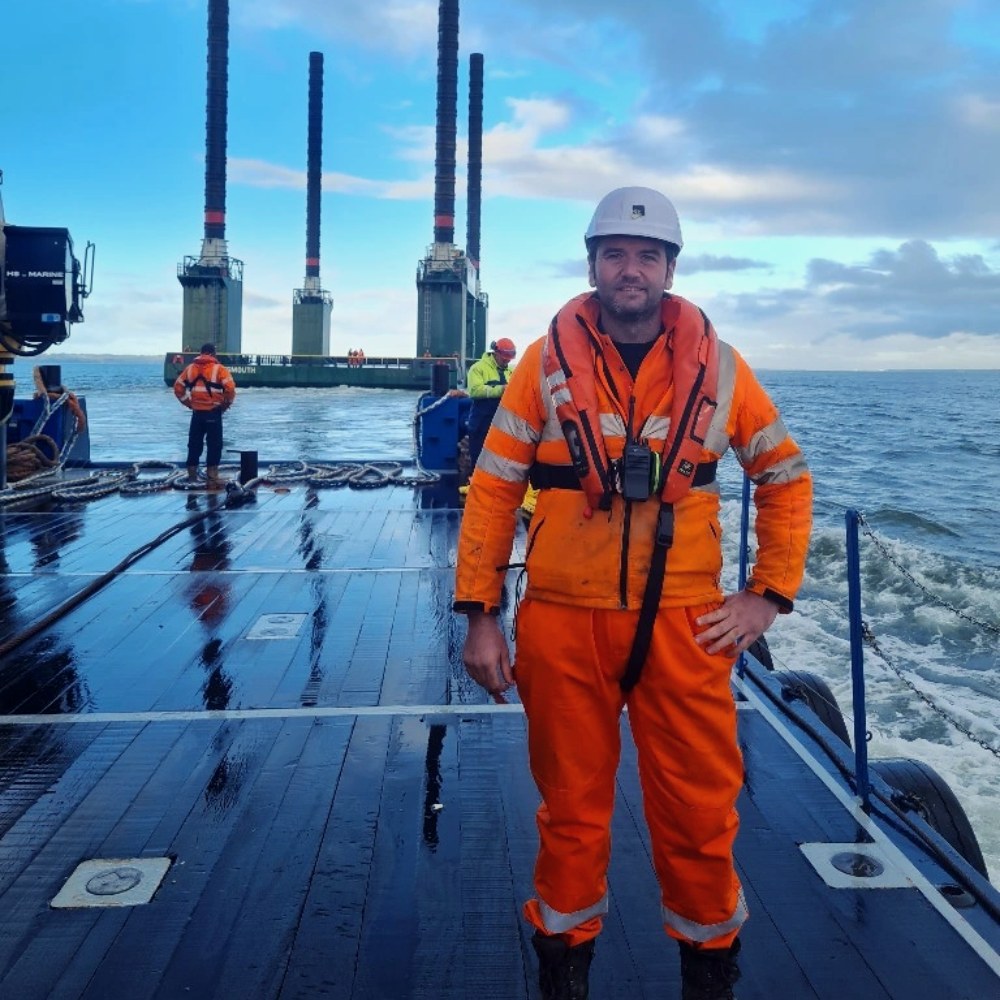 Engineer in orange coveralls and a lifejacket on a workboat deck, with a jack-up barge behind