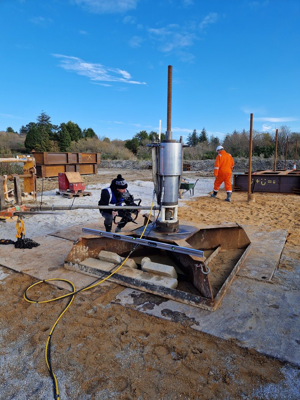 Drilling equipment on a base frame with an anchor bar rising from it, engineers alongside