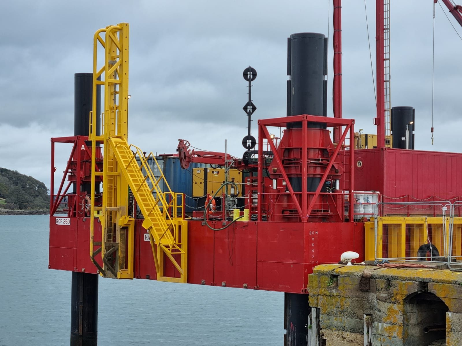 Red jack-up barge with yellow access stairs and drilling equipment, moored at a stone quay
