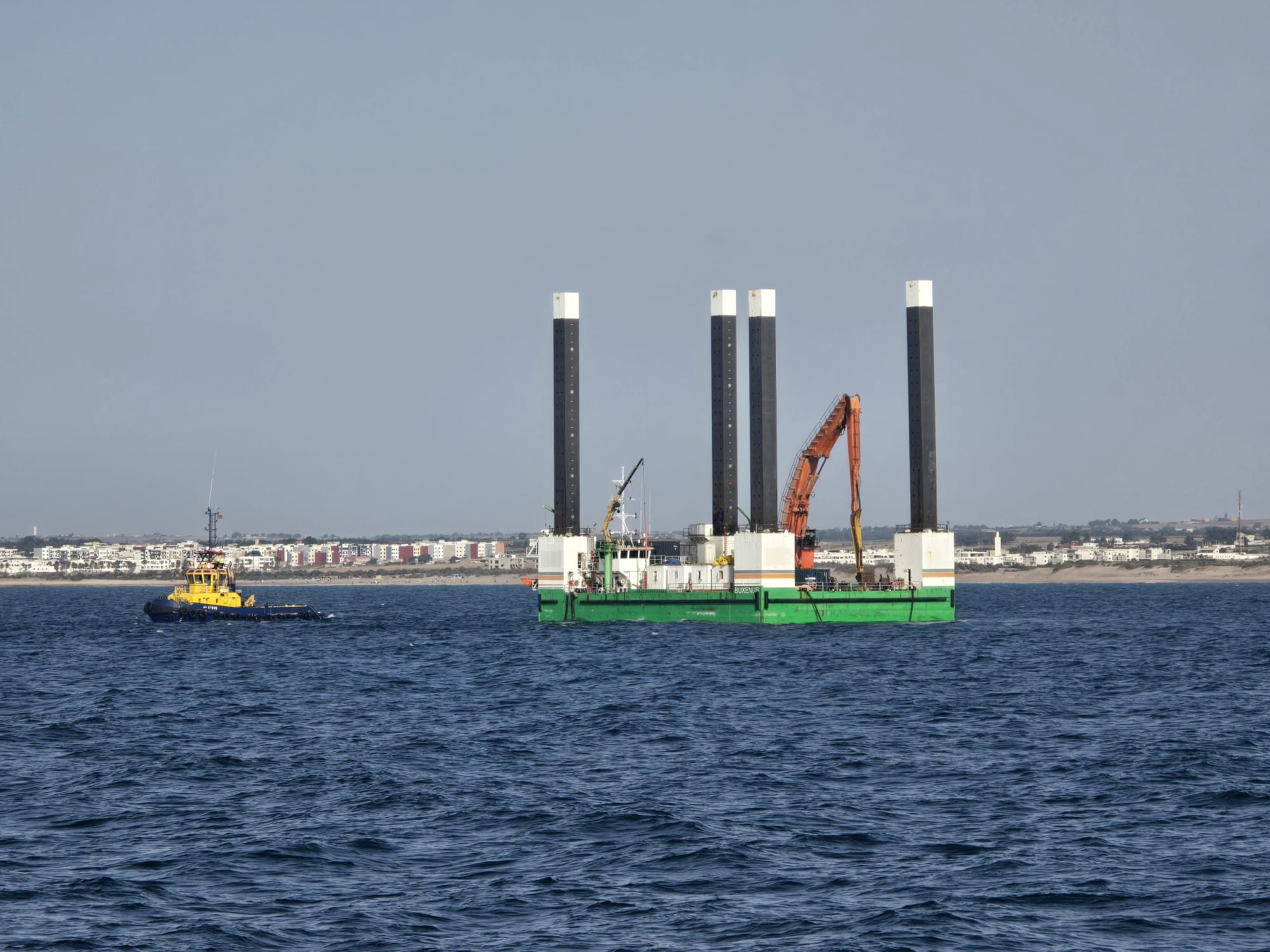Green jack-up barge with four legs raised, working off a coastline with a tug nearby