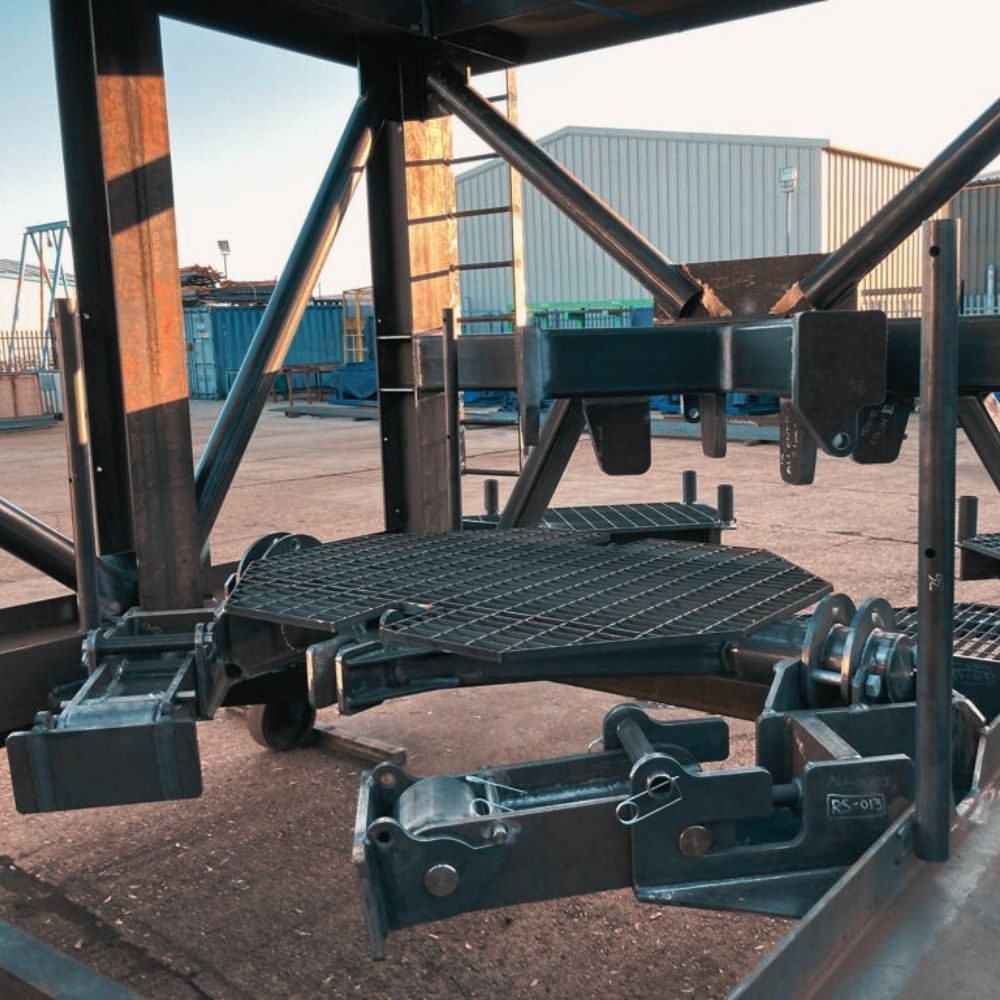 Dark steel guide frame with hinged latches, seen from beneath in a fabrication yard