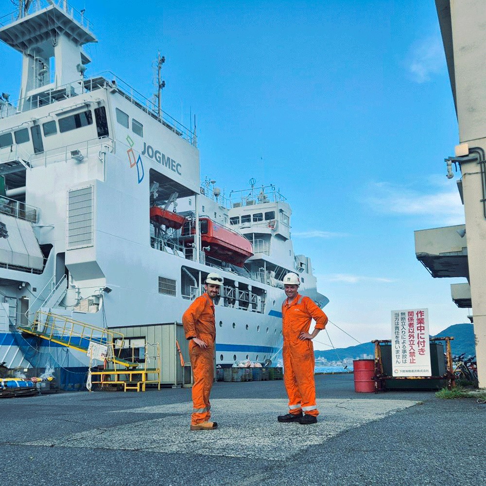 Two engineers in orange coveralls on a quay beside a white research vessel