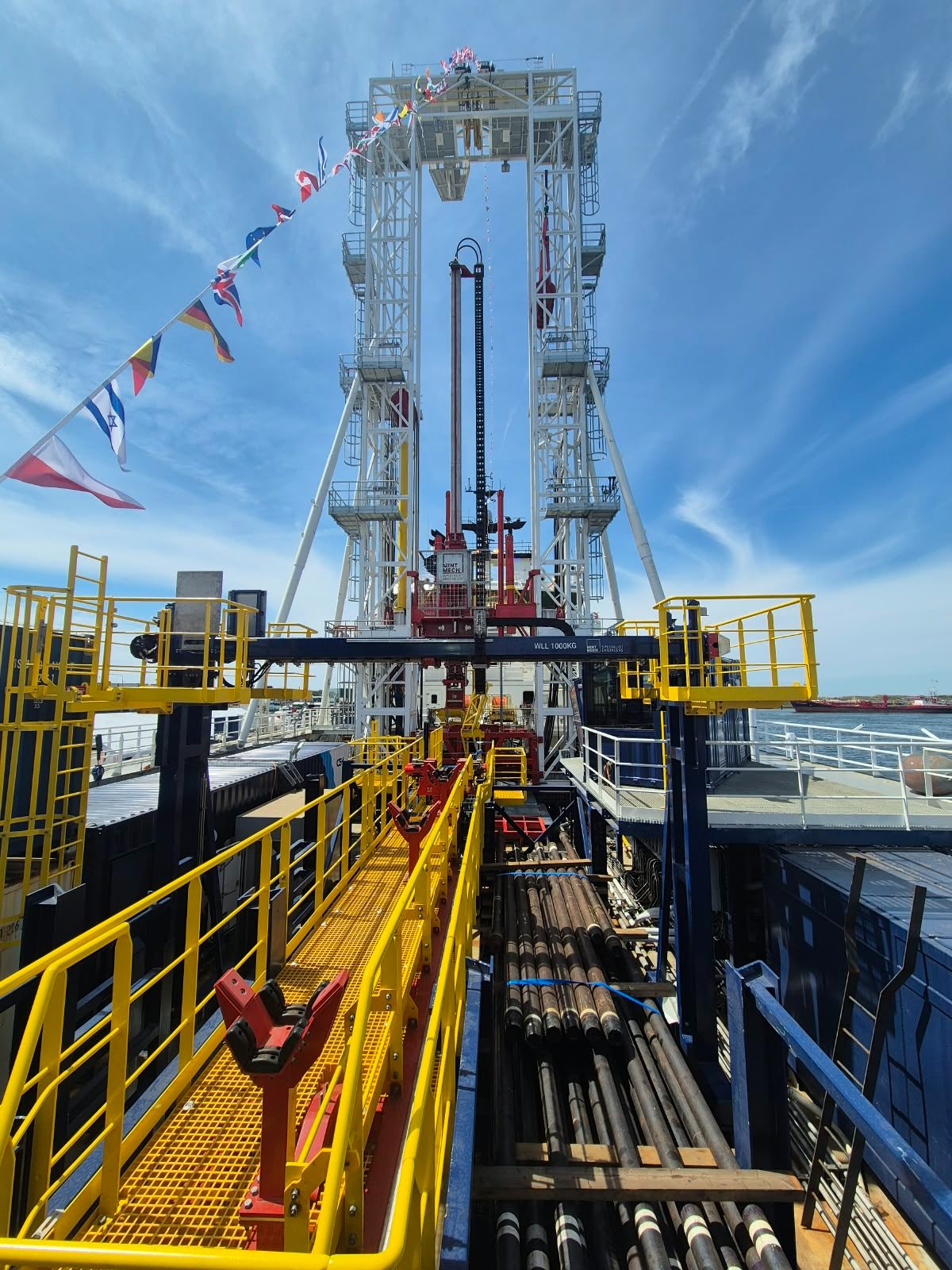 Geotechnical drilling derrick dressed with flags above a pipe rack and yellow walkway