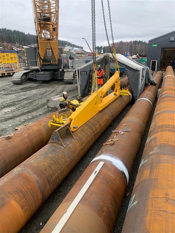 Crane lowering a yellow lifting tool onto large steel tubular piles in a yard